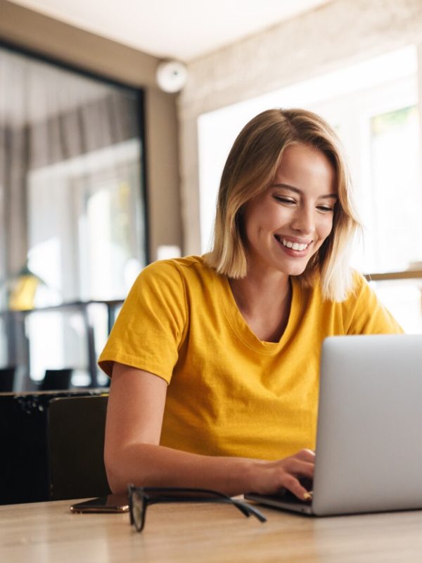 Photo of joyful nice woman using laptop and smiling while sitting Photo of joyful nice woman using laptop and smiling while sitting at table in open-plan office